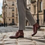 Kobieta w bordowych oksfordach i wełnianych spodniach idąca po słonecznym bruku w Paryżu. Zbliżenie na buty i detale tkanin.